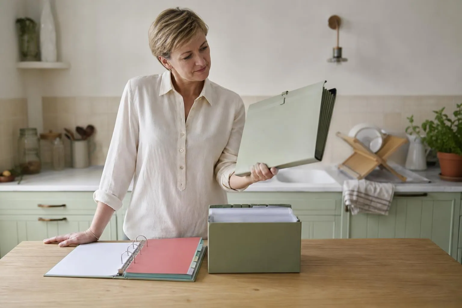Assistante maternelle d’environ quarante sept ans debout devant une table où sont alignés un classeur à intercalaires ouvert, une boîte de pochettes par enfant et un porte documents, dont elle soulève un exemplaire.